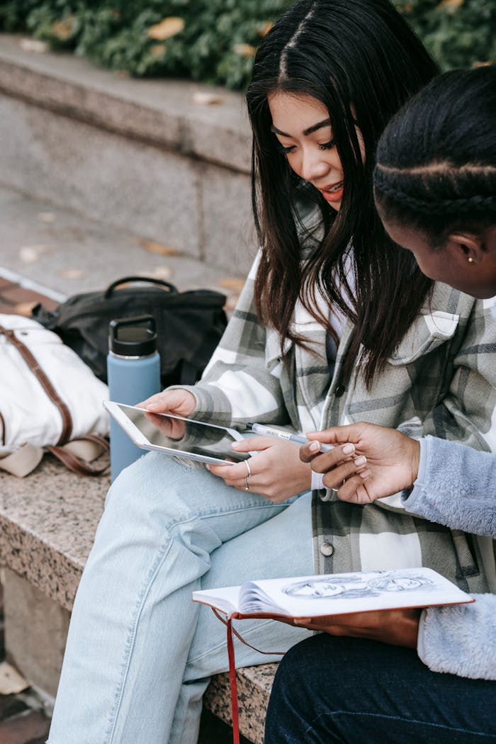 portfolio-06 Crop young Asian woman showing cellphone with black screen to unrecognizable ethnic female friend while talking on city bench